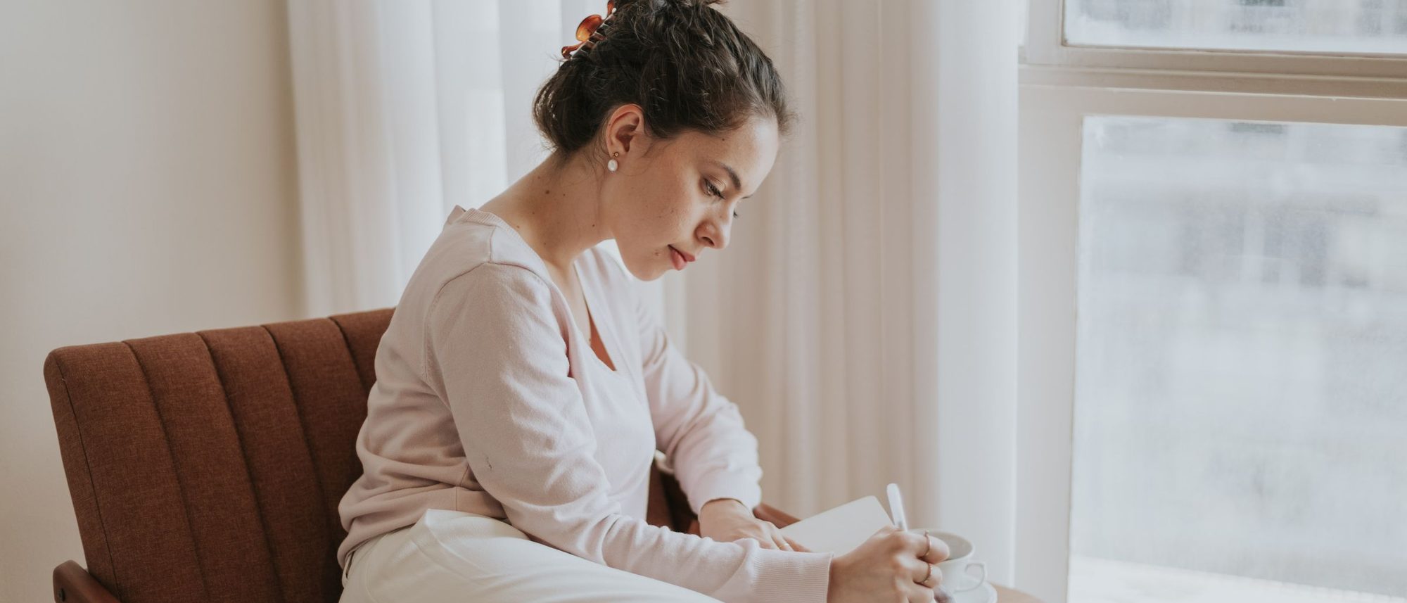 A woman writing in a journal in a comfortable chair by a sunlit window
